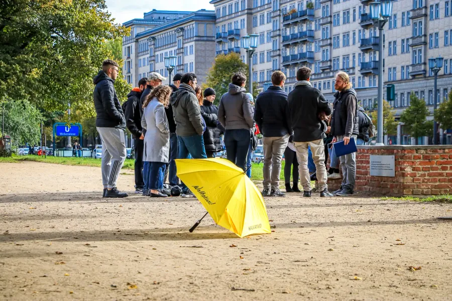 A group of people listening to a guide near apartment buildings in Berlin. A yellow Walkative umbrella rests on the ground.
