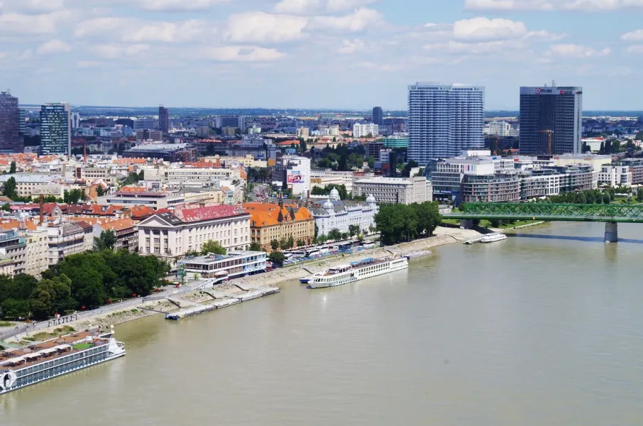 Aerial view of Bratislava cityscape along the Danube River, featuring modern skyscrapers, historic buildings, and a green arc