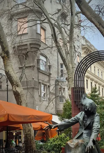 Bronze statue of Franz Liszt sitting, with bare trees, an orange cafe umbrella, and buildings in the background on a winte...
