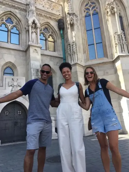 Three smiling people stand arm-in-arm in front of the ornate, light-colored Matthias Church with Gothic windows.