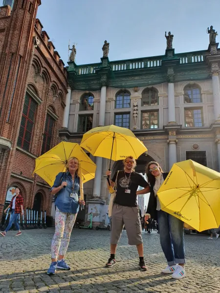 Three smiling Walkative guides hold yellow umbrellas on a cobblestone street in front of Gdańsk's historic Green Gate buil...