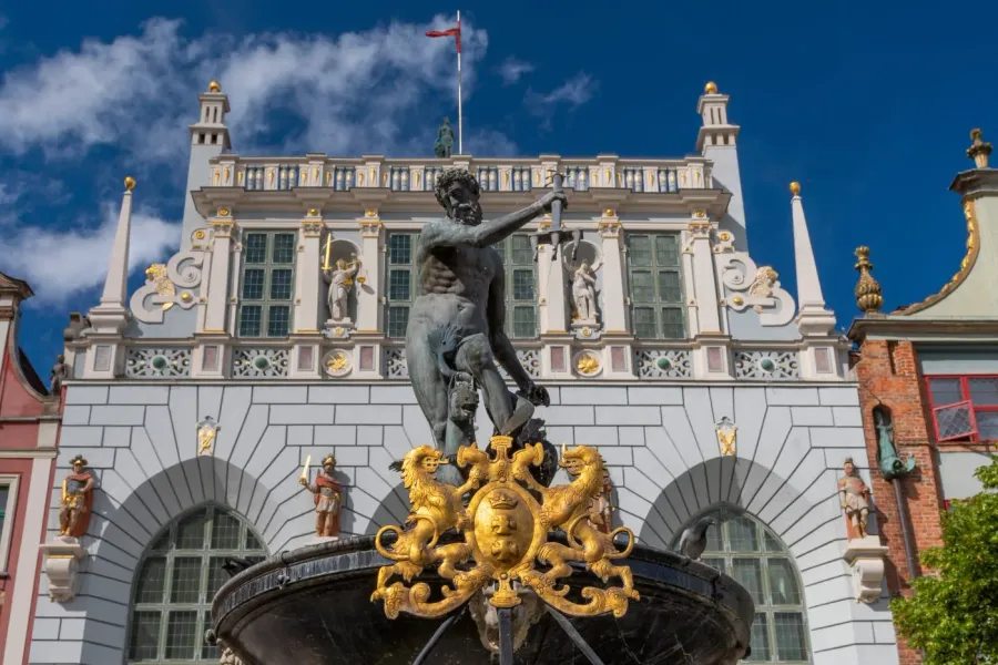 Bronzestatue des Neptun mit Dreizack auf einem Brunnen vor einem prächtigen, reich verzierten Gebäude bei blauem Himmel.