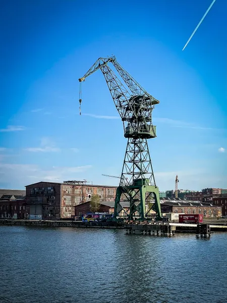 Large green gantry crane standing over the Motława River, with historic shipyard buildings under a clear blue sky in Gdańsk.