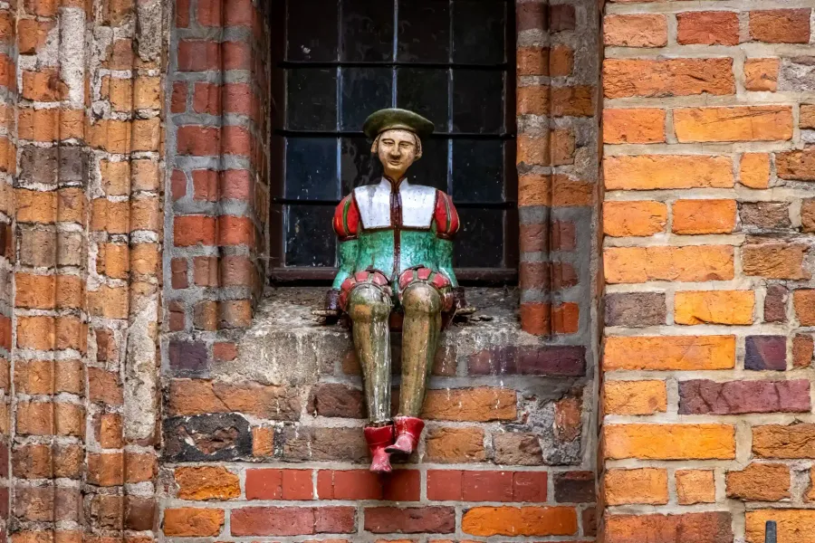 Bunte Holzfigur eines Mannes mit grüner Baskenmütze und mittelalterlicher Kleidung, sitzend auf einer Ziegel-Fensterbank e...