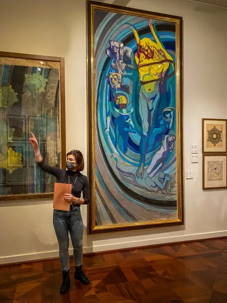 A tour guide wearing a face mask stands in an art gallery, pointing towards a large colourful painting on the wall.