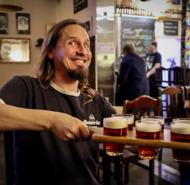 Man with long hair and beard holding a beer flight with five small glasses of amber beer in a pub.