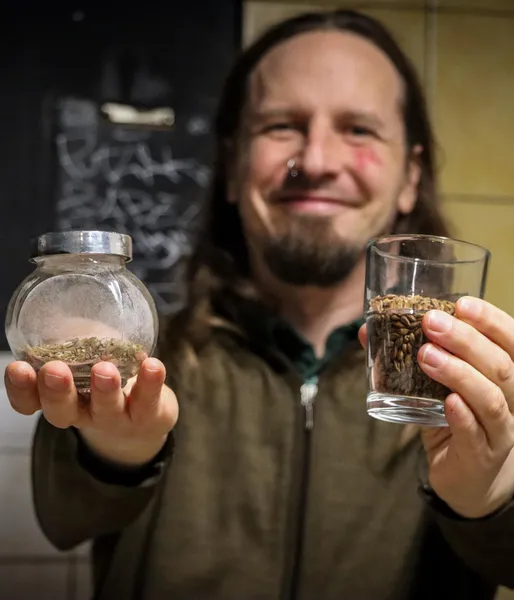 Ein Mann mit langen Haaren hält ein Glas mit Gerstenmalz und ein kleines Glas mit getrocknetem Hopfen in die Kamera.