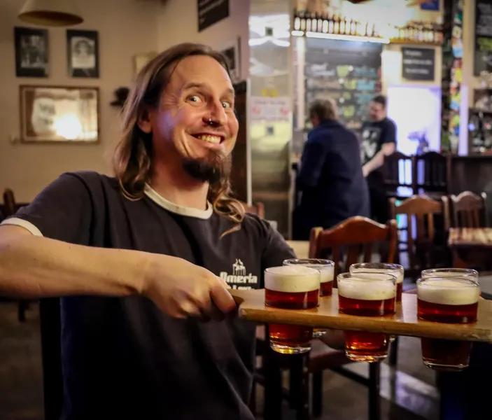 A smiling man with long hair and a nose ring holds a wooden flight with six small glasses of beer in a pub.