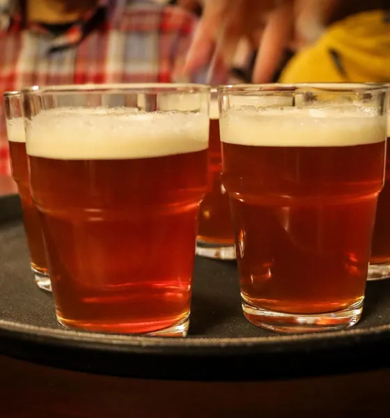 Several glasses of amber-colored beer with foam heads are arranged on a dark serving tray in a pub.