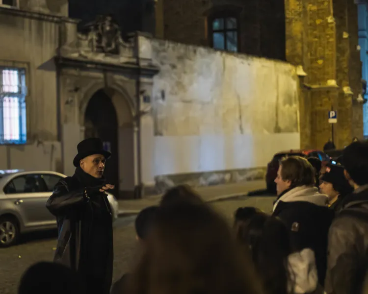 Tour guide in top hat and long coat speaking to a group at night on a cobblestone street in Krakow. Historic buildings are...