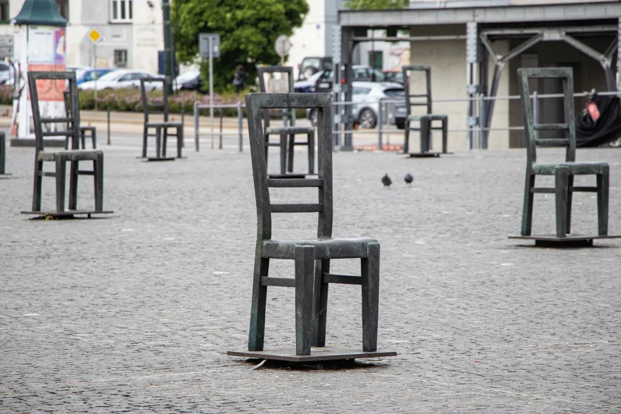 Esculturas de sillas de metal llenan una plaza adoquinada, con dos palomas visibles y edificios al fondo.