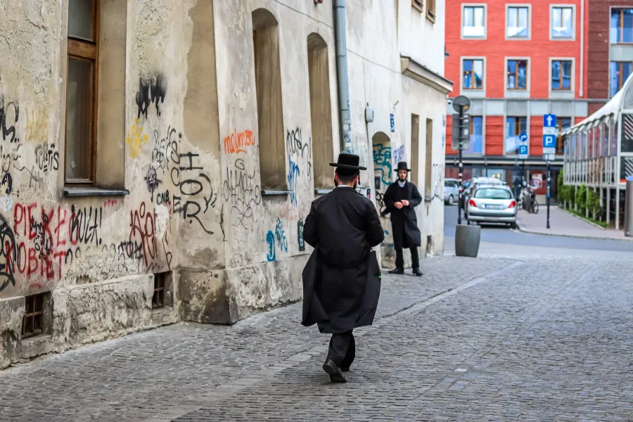 Two Orthodox Jewish men on a cobblestone street in Krakow's Kazimierz district, near a wall covered in graffiti.