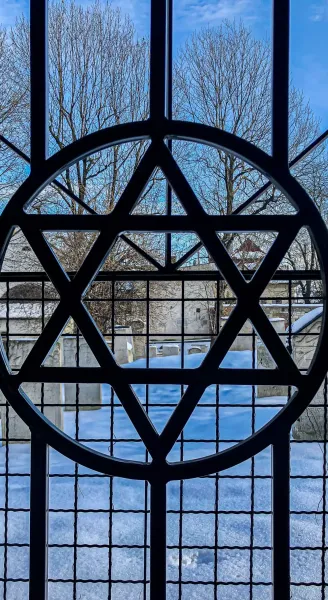Blick durch ein Fenster mit einem Davidstern-Gitter auf einen schneebedeckten Friedhof im Winter.