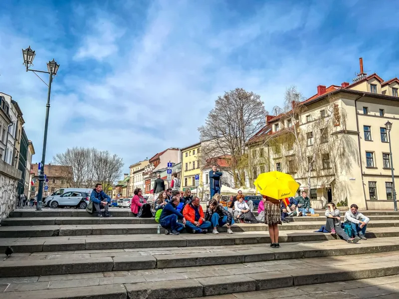 Eine Reisegruppe sitzt auf breiten Steinstufen auf einem Stadtplatz in Krakau und lauscht einem Führer mit gelbem Schirm.