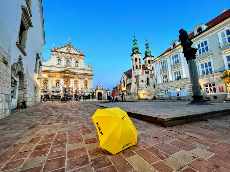 Żółty parasol Walkative na bruku placu w Krakowie, z zabytkowymi kościołami w tle o zmierzchu.