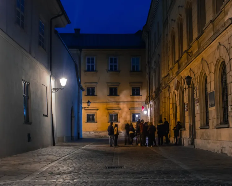 A tour group stands on a narrow, lamplit cobblestone street in Krakow at night, surrounded by historic buildings.