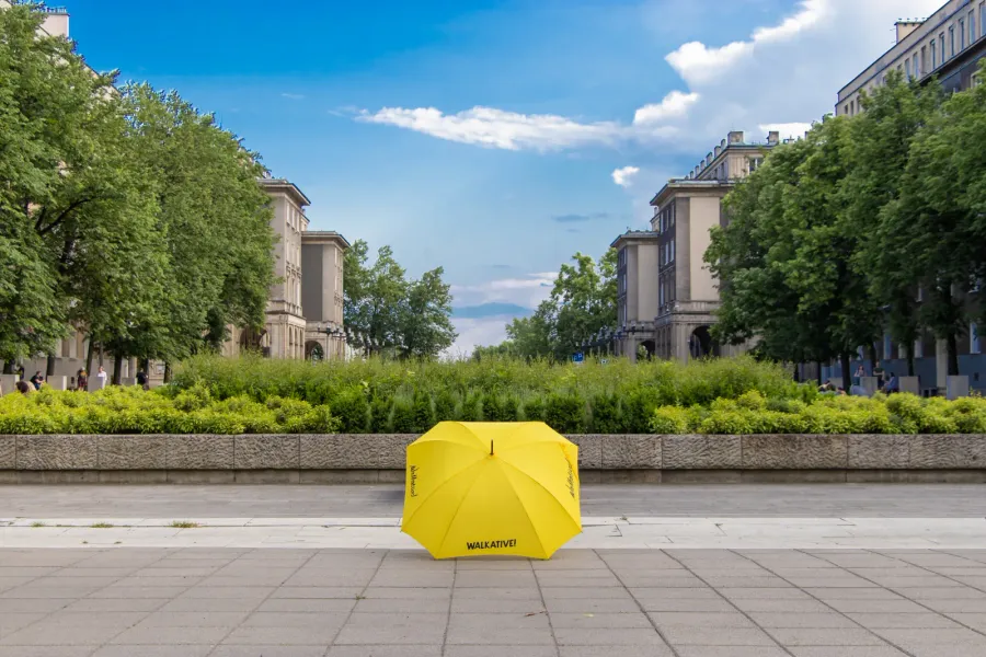 A yellow Walkative umbrella on the pavement of a large square in Nowa Huta, with trees and socialist-realist buildings.