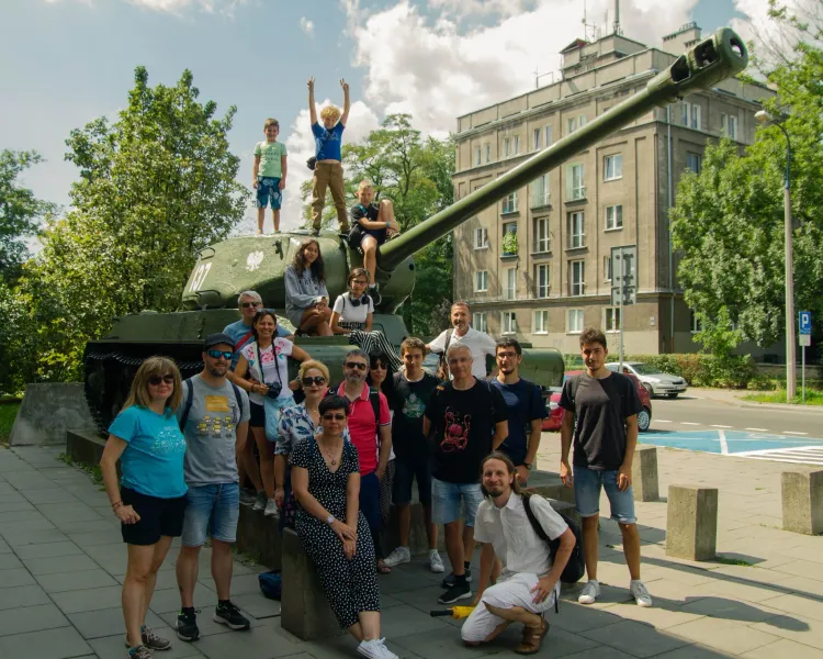 A tour group of adults and children posing for a photo on and around a green IS-2 tank monument in Krakow's Nowa Huta.