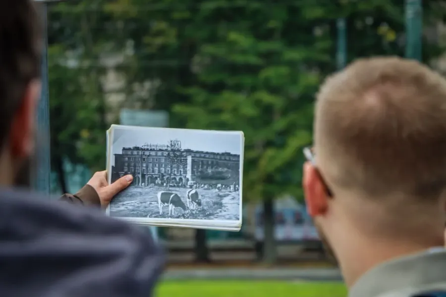 A tour guide shows a visitor a historical black and white photo of a building with cows grazing in front of it in Nowa Huta.