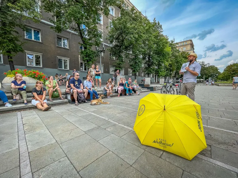 A tour guide speaks to a group of people sitting on steps in a city square, with a yellow Walkative umbrella in front.