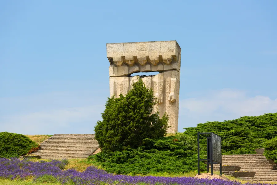 Ein großes, abstraktes steinernes Denkmal steht vor einem klaren blauen Himmel, umgeben von grünen Sträuchern, Steintreppe...