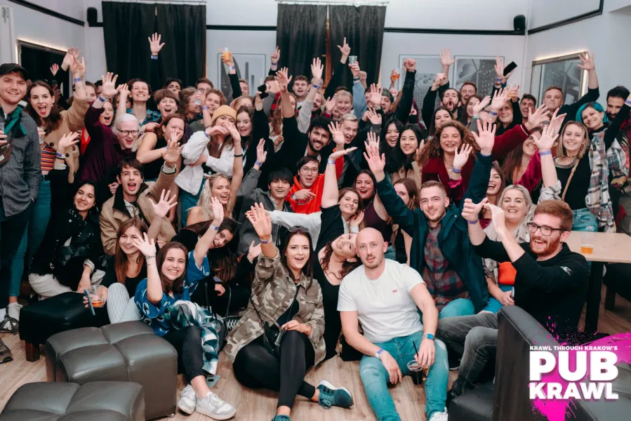 Duża grupa młodych ludzi uśmiecha się i macha do kamery w barze podczas pub crawl w Krakowie.