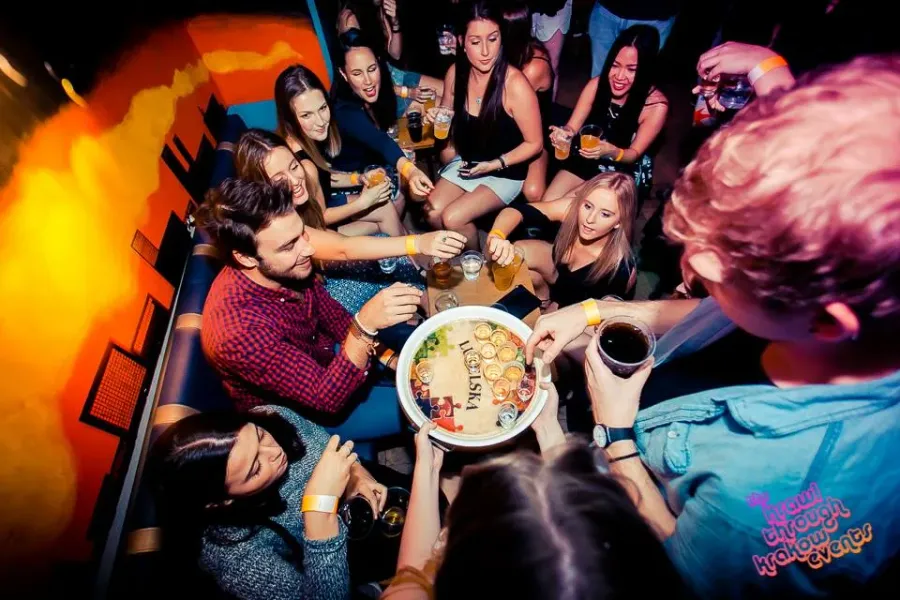 Grupa młodych ludzi w barze, dzieląca się okrągłą tacą z shotami podczas imprezy typu pub crawl.