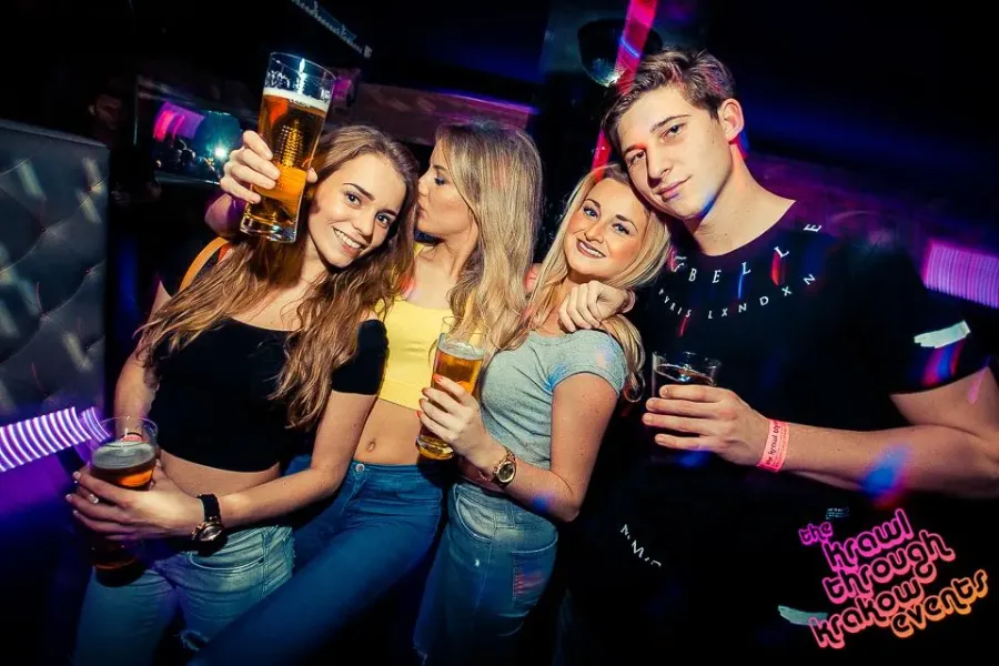 Grupa czworga młodych ludzi uśmiecha się i trzyma szklanki z piwem w słabo oświetlonym klubie nocnym.