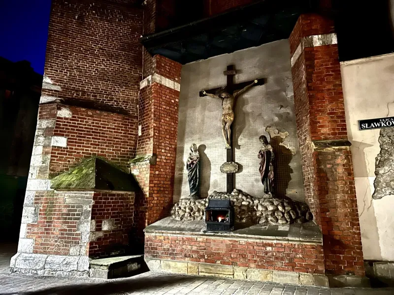 A streetside shrine with a crucifix set into a brick wall on Sławkowska Street in Krakow at night.