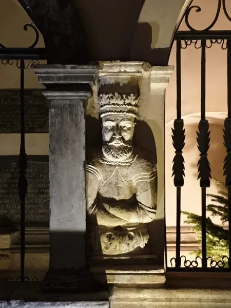 A weathered stone sculpture of a bearded king with a crown, built into a pillar and illuminated from below at night.