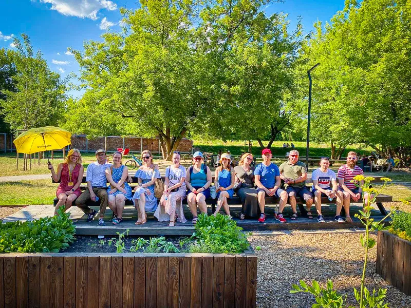 Ein Tourguide mit gelbem Regenschirm sitzt mit einer Gruppe von Touristen auf einer langen Bank in einem sonnigen Park in ...
