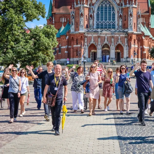 Un guía con un paraguas amarillo lidera a un grupo que saluda a la cámara frente a una gran iglesia de ladrillo en Cracovia.