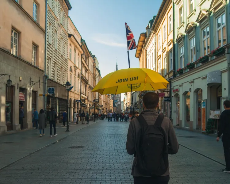 Przewodnik trzyma żółty parasol na zabytkowej, brukowanej ulicy w Krakowie, prowadząc grupę w stronę wieży kościelnej.