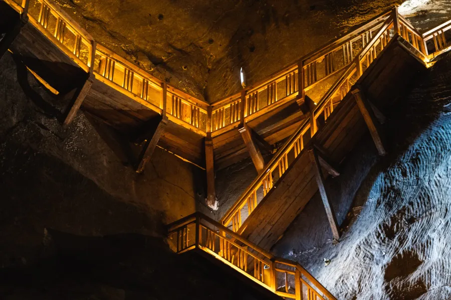 A large wooden staircase structure, lit with warm yellow light, built against the dark, rough walls of a salt mine chamber.