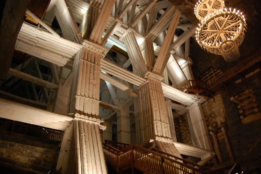 Massive wooden support beams and pillars inside the Wieliczka Salt Mine, with two large salt crystal chandeliers hanging.