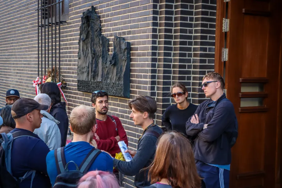 Ein Reiseführer erklärt einer Gruppe von Reisenden etwas vor einer Gedenktafel an einem Backsteingebäude in Krakau.
