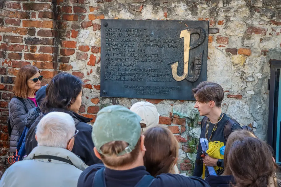 Un guía turístico explica a un grupo de turistas una placa histórica con el Símbolo de la Lucha de Polonia en una pared.