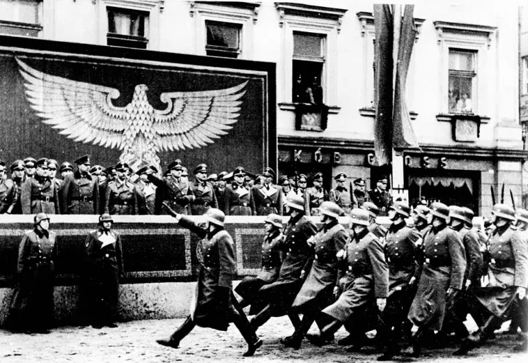 Schwarz-Weiß-Foto einer NS-Militärparade in Krakau. Soldaten marschieren an einer Tribüne mit Offizieren und einem großen ...
