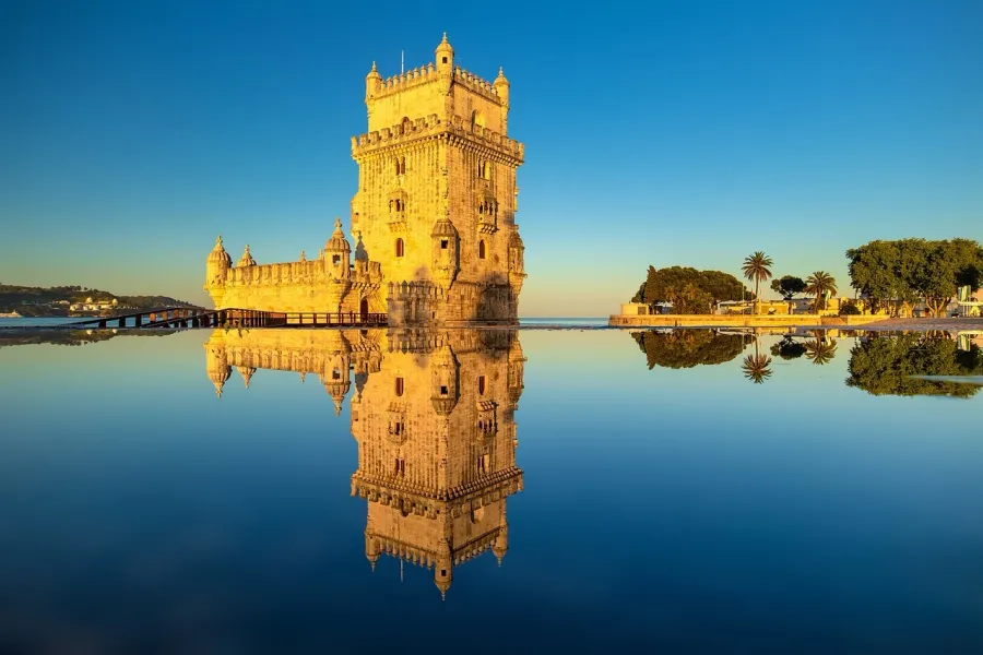 Belém Tower in Lisbon illuminated by golden light, perfectly reflected in the calm water against a clear blue sky.