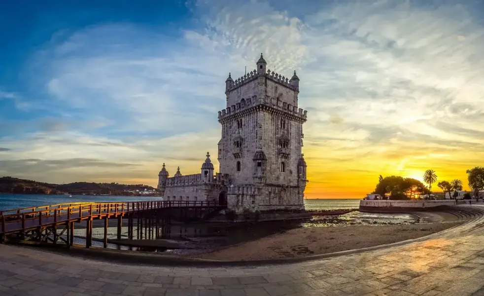 Historischer Turm von Belém in Lissabon bei Sonnenuntergang, mit Holzsteg über dem Tejo und orange-blauem Himmel.