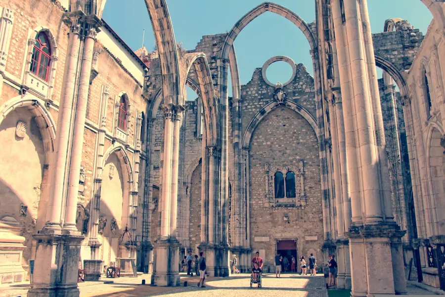 Tourists explore the roofless Gothic arches and stone ruins of the Carmo Convent in Lisbon under a clear blue sky.