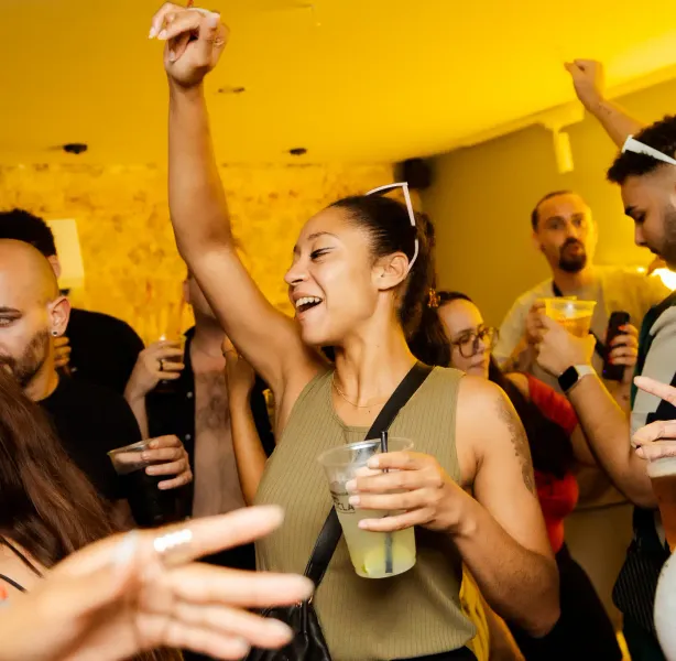 Smiling woman with drink, arm raised, dancing in a lively group at a Lisbon nightlife spot. Yellow light, blurred people.