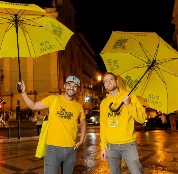 Two men in yellow 'The Lisbon Pubcrawl' apparel holding yellow umbrellas, standing on a wet street at night in Lisbon.