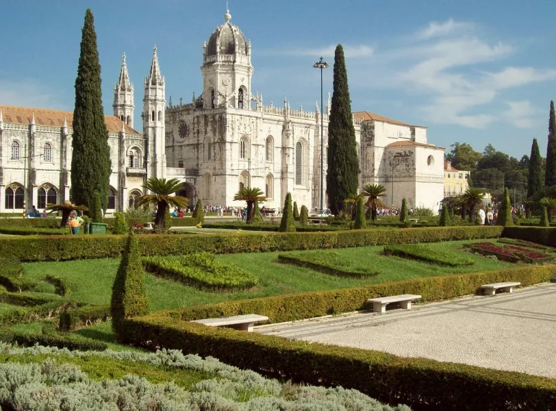 Jerónimos-Kloster in Lissabon mit gepflegten grünen Gärten, Zypressen und Besuchern an einem sonnigen Tag.