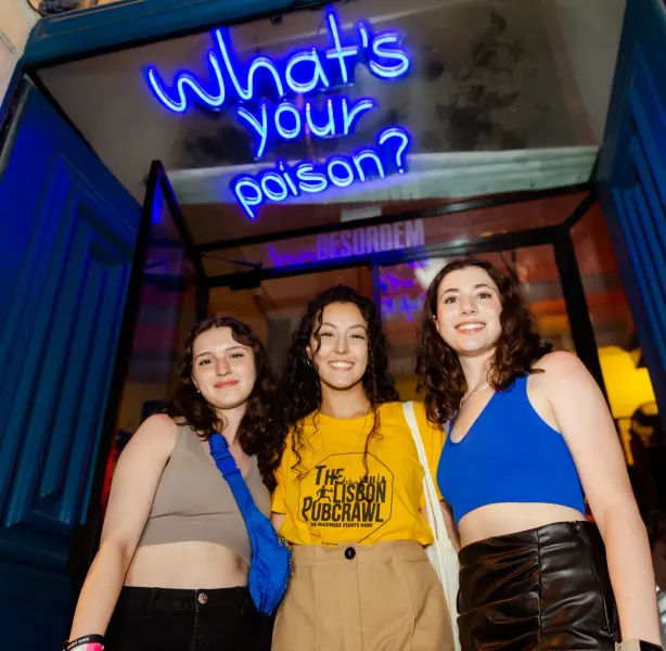 Three young women smiling in front of a blue neon sign that reads 'What's your poison?' above a blue door frame.
