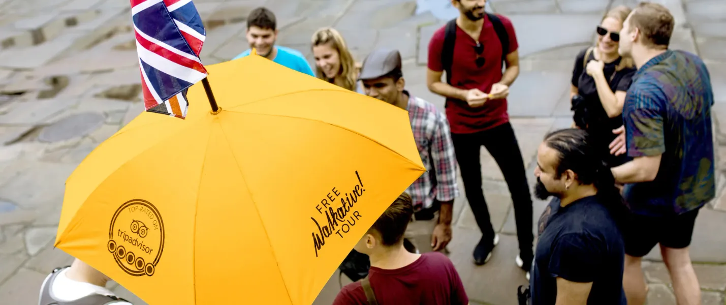 Eine vielfältige Gruppe von Menschen hört einem Reiseleiter zu, der einen gelben Walkative-Regenschirm mit kleiner Union Jack