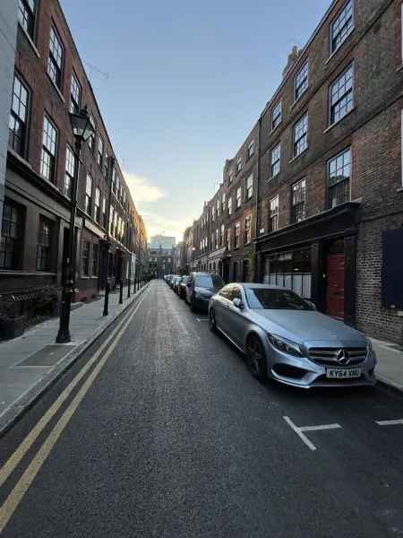 A narrow London street with cars parked between historic brick buildings at sunset.
