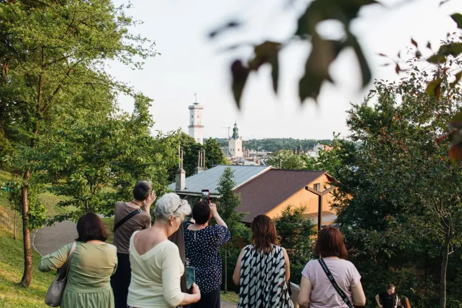 Group of people on a hill overlooking Lviv cityscape with City Hall tower and a church dome visible through trees.