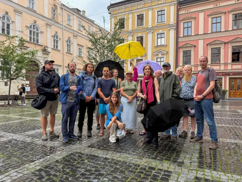 A group of about 15 adults stands on a wet cobblestone square in Lviv. Several hold umbrellas, with colorful old buildings...