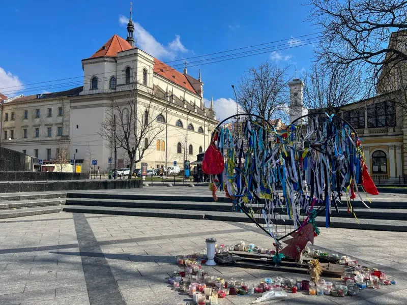 Monumento en forma de corazón con cintas azules y amarillas y velas en una plaza de Leópolis, con un edificio histórico de...